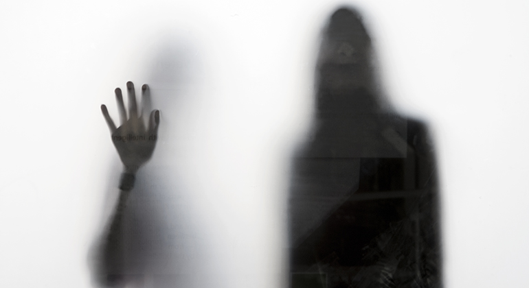 Svartvitt foto. En person har lagt sin hand på en dimmig ruta. I bakgrunden en svart siluett av en människa som ger ett hotfullt intryck. 
