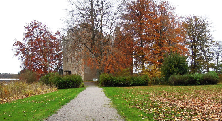 En bred grusgång leder fram till ruinen som med sina höga murar skymtar bakom de höstfärgade träden.