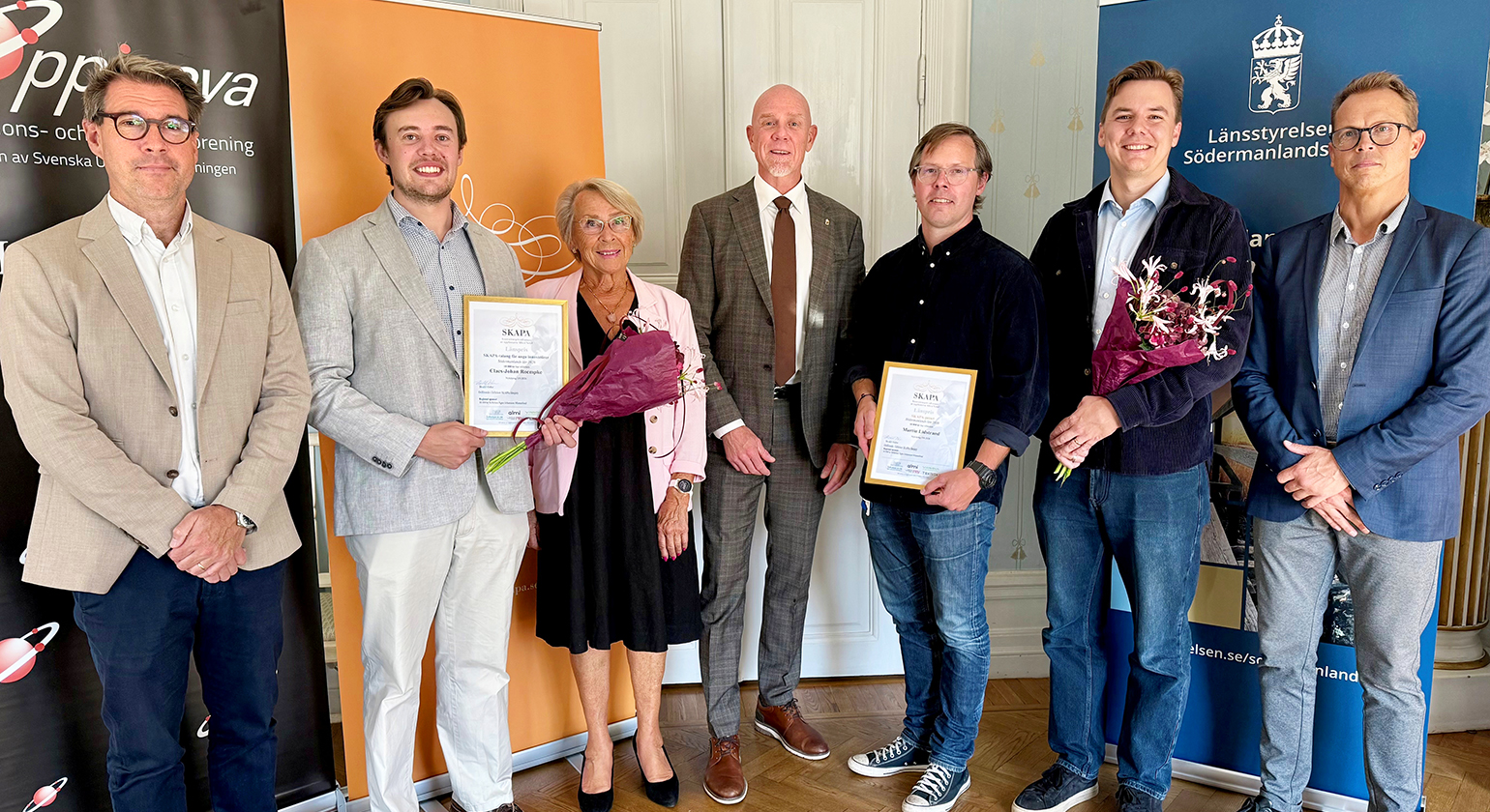 Sju människor står bredvid varandra i en sal. Diplom och blombuketter visas upp.