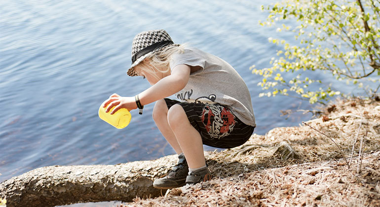 Ett barn klädd i tröja, shorts och hatt sitter på huk på stranden.
