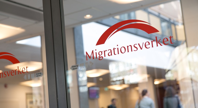 Glasdörr med Migrationsverkets logotyp. Innför syns en taklampa och människor i grupp.