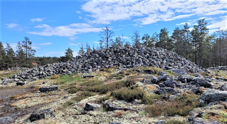 Öppen stenbunden mark med en del gräs. I bakgrunden syns en tät barrskog. På den öppna marken ligger ett stort stenröse uppbyggt av famnstora stenar. 
