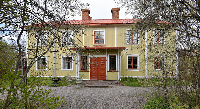 Huset har en gul-grön färg och ett orange plåttak. De orangea detaljerna återfinns vid fönstren och dörren.