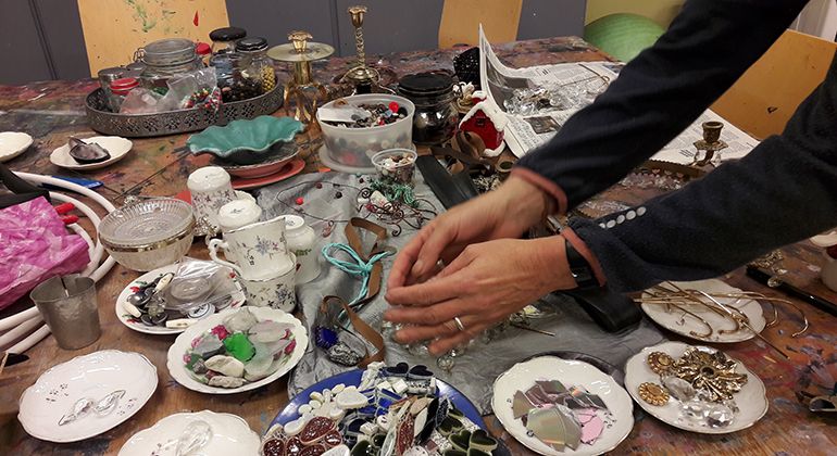 Ett par händer som sträcker sig över ett färgfläckat och nött bord fullt av pärlor, stenar och annat pysselmaterial.