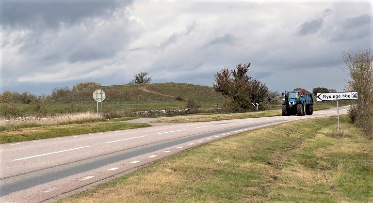 En bild på en landsväg som leder diagonalt in i bilden. En traktor är på väg mot betraktaren. Vid vägkanten står en skylt med pil och texten Mysinge hög. Skylten pekar mot en hög gräsklädd kulle som finns på andra sidan vägen och bakom en mindre stenmur. 