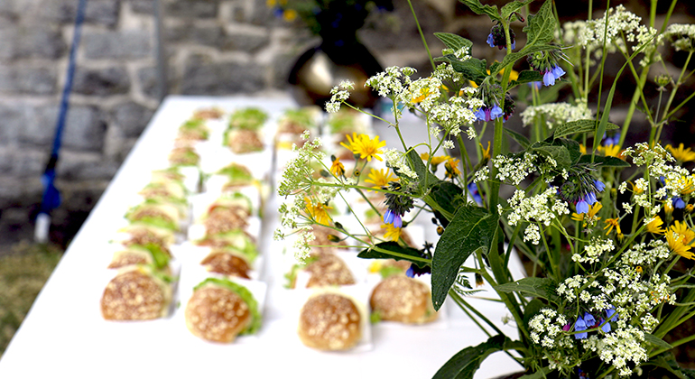 Ett bord med vit duk uppdukat med smörgåsar. I förgrunden syns en bukett blommor i gult, blått och vitt.