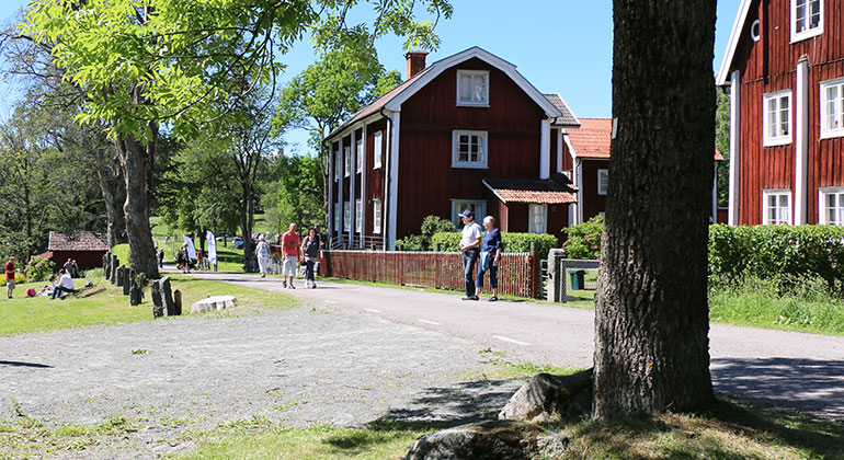 Människor som går på en väg, röda hus med vita knutar, en trädstam längst fram i bilden.