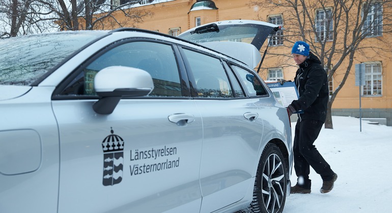 En silverfägad bil med Länsstyrelsen Västernorrlands logotyp i svart på förardörren. I bakgrunden håller en person på att lasta in något i bagageluckan.