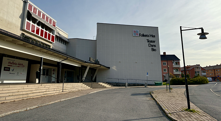 Avlång byggnad med en högre byggnadskropp ställd i vinkel. I hörnet på byggnaden står det på fyra rader: Folkets hus, Teater, Dans och Bio. I bakgrunden en gång- och cykelväg, lägenhetshus i två till tre våningar.