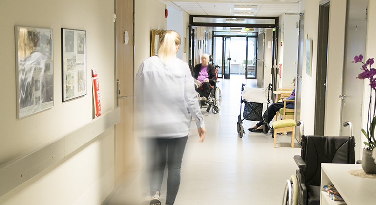 Kvinna som går genom en korridor på ett äldreboende. Längre ner i korridoren sitter en äldre dam i en rullstol.