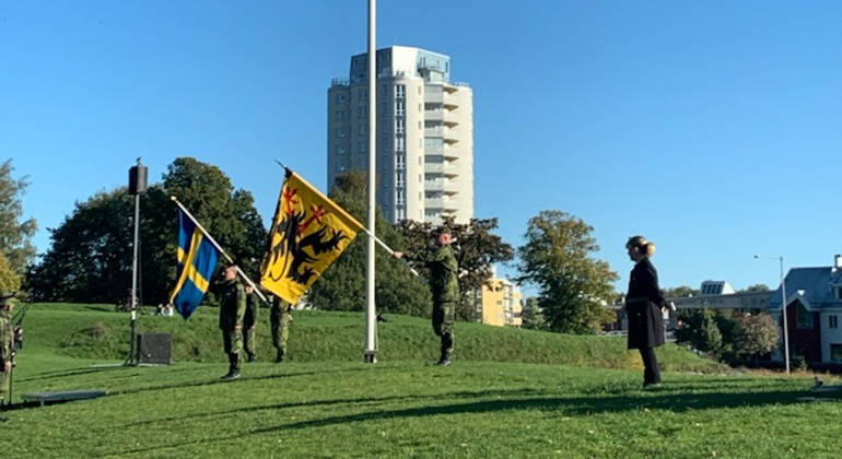 Landshövdingen deltar i avslutning av militärövning. En militärklädd man håller en fana med länsvapnet och en håller i en svensk fana.