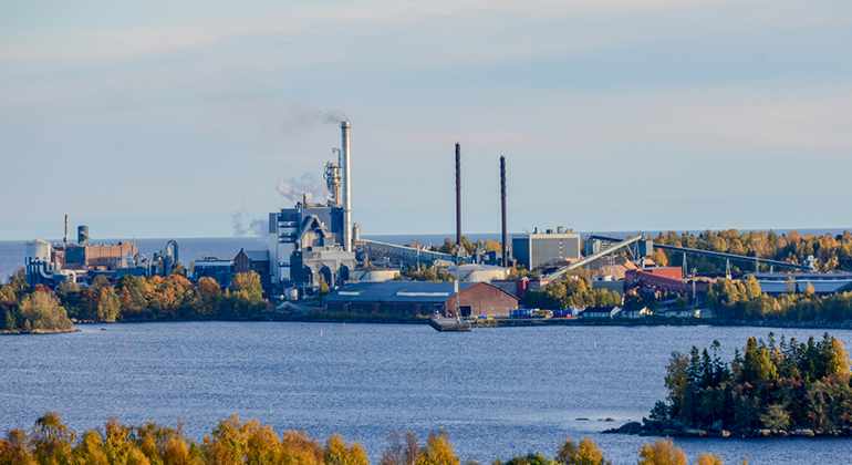 En fabrik som ligger vid vattnet. Rök kommer upp ur skorstenar. Fabriken består av flera byggnader.