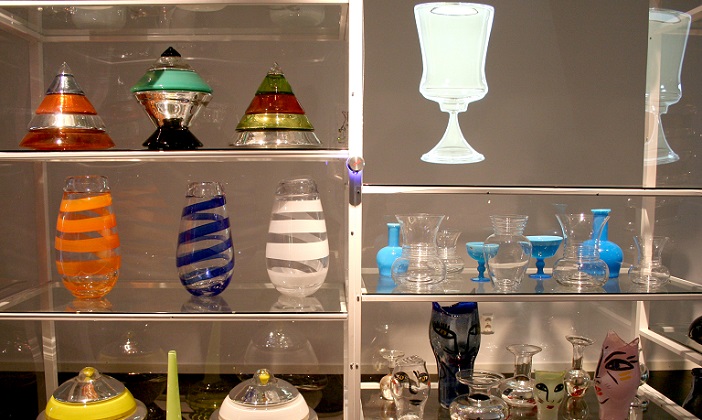 Flera belysta hyllor på vilka står glasföremål i olika former och färger som vaser och skålar.