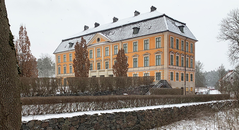 Nynäs slott, en stor byggnad i färgerna gult och beige. Framför byggnaden finns träd, buskar och stenmurar. 