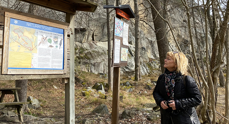 Landshövding Beatrice Ask står ute i naturen och tittar på en skylt för naturreservatet Simonberget.