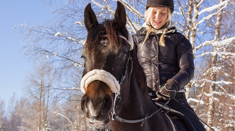 Flicka rider på en brun ponny med vit bläs som har frost i morrhåren. Det är vinter ute och flickan har varma kläder och ridhjälm.