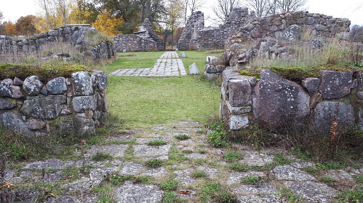 En rektangulär ruin sedd inifrån med väggar av murade naturstenar av varierande höjd. En gång med plattor syns i mitten av ruinen, i övrigt består marken av gräs. Några träd och buskar i bakgrunden, varav några gula i höstfärger.