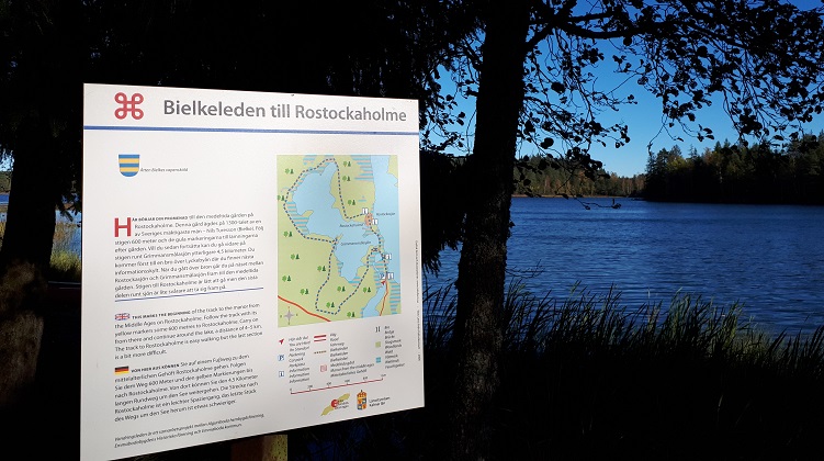 En solbelyst informationstavla med text och karta som det står Bielkeleden till Rostockaholme på. I bakgrunden en sjö med mycket blått vatten.