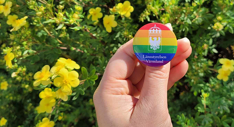Hand som håller i regnbågsfärgad knapp med Länsstyrelsen Värmlands logotyp.