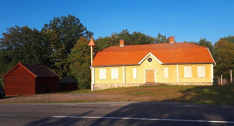 En stor gul träbyggnad rött tegeltak. Ett litet klocktorn finns vid ena gavel samt en mindre fristående rödmålad träbyggnad bredvid.