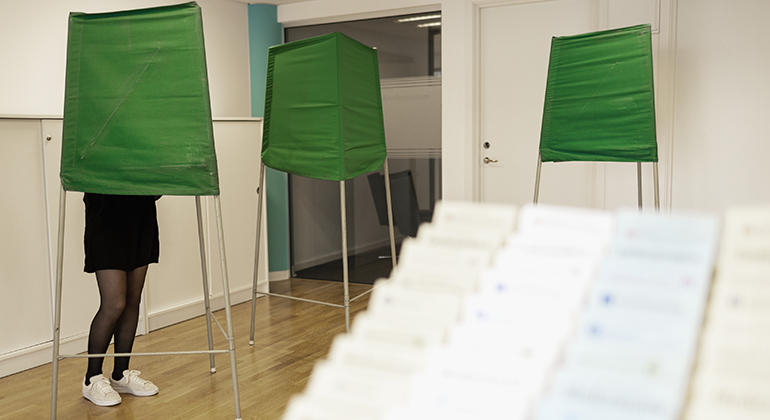 Foto på kvinna som står och röstar i ett grönt valbås i en vallokal.