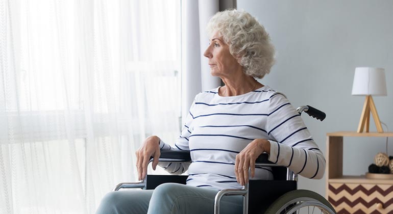 Äldre kvinna i rullstol tittar ut genom ett fönster. 