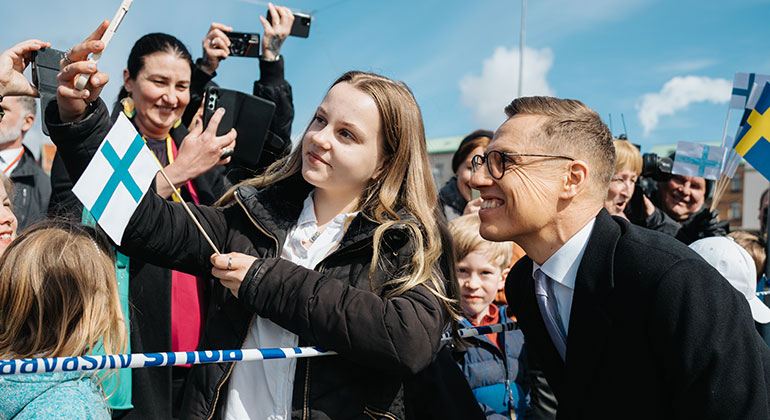 I förgrunden en flicka med en finsk flagga som fotar sig själv med president Alexander Stubb. I bakgrunden människor som fotar. 