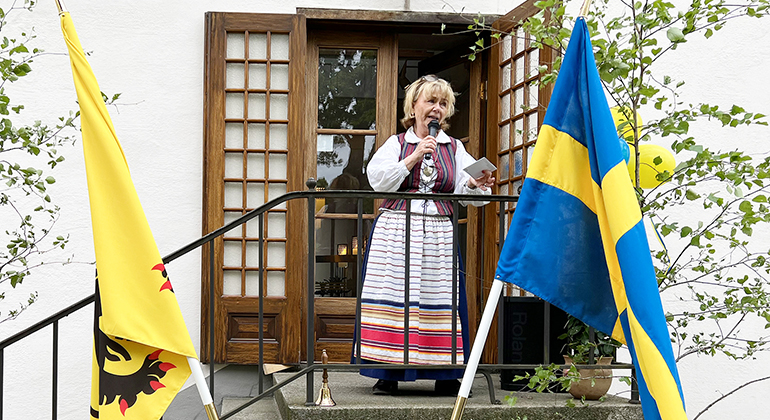 Beatrice Ask står och pratar i mikrofon på en trapp vid residenset, framför hänger länsstyrelsens flagga och den svenska flaggan.