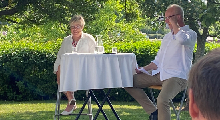 En kvinna och en man sitter vid ett bord ute i en trädgård och samtalar.