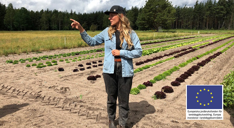 Foto av en kvinna som står på en fält med växande grönsaker i bakgrunden. 