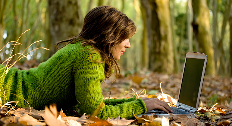 En mörkhårig kvinna i grön polotröja ligger på mage på marken i skogen. Hon har en bärbar dator framför sig.