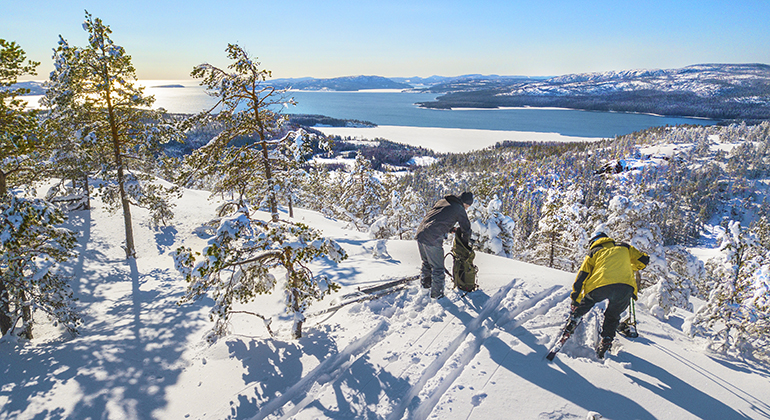 Snöigt landskap med ungtallar som vajar i vinden. Två personer står redo att sätta på sig skidor och susa ner för en brant. De har utsikt över havet där de står.