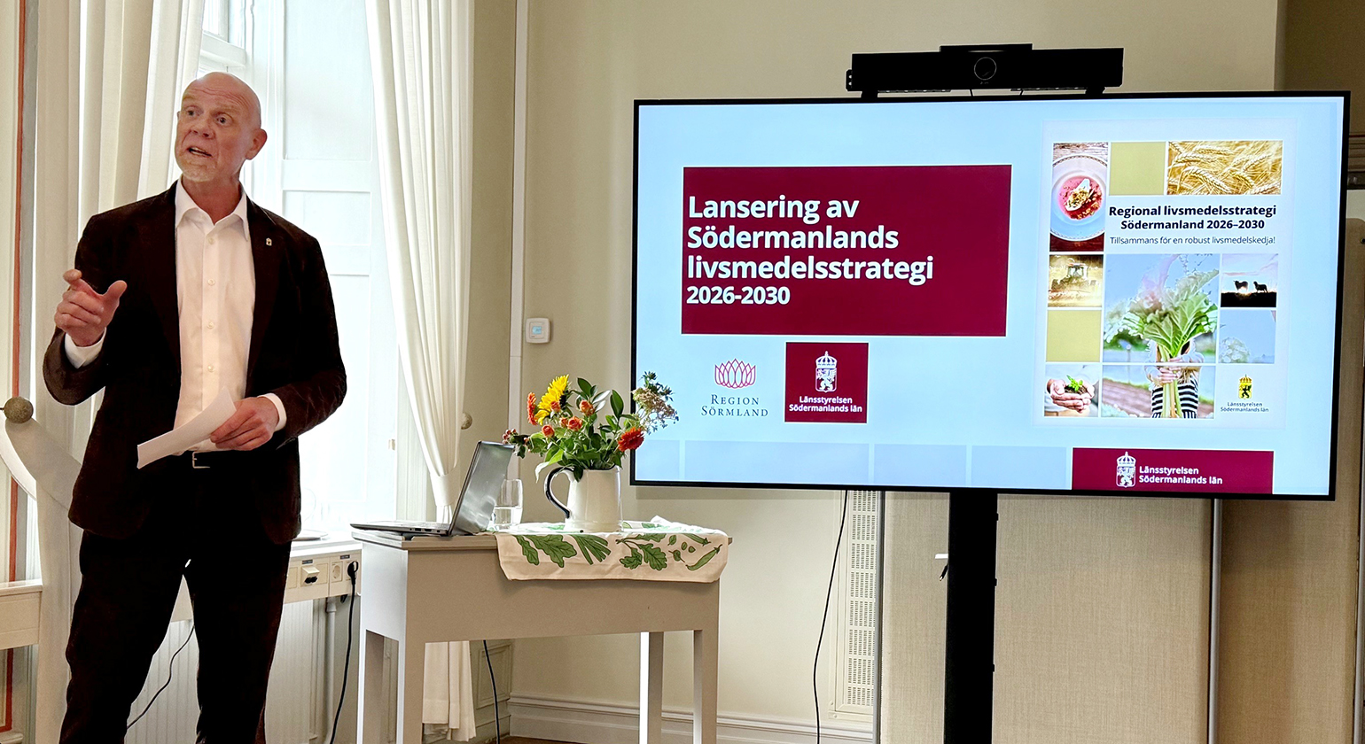 Robert Andrén står i en sal och pratar, bredvid honom syns en stor bildskärm med presentation av livsmedelsstrategin.