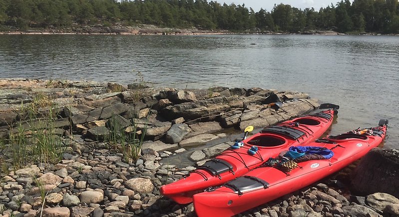 Två kajaker ligger uppdragna på en stenig strand en mulen sommardag. I bakgrunden syns lugnt vatten och skogsklädda öar.