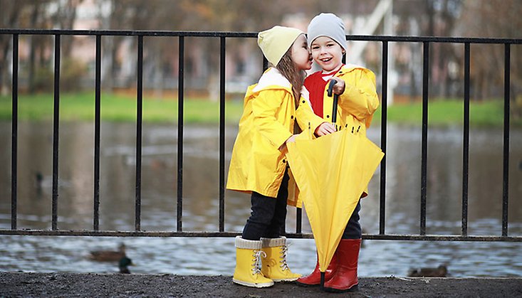 Två barn i gula regnkläder, gummistövlar och med ett paraply. De står framför ett staket med vatten i bakgrunden.