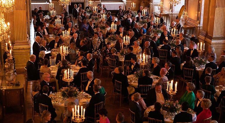 Översiktsbild över en middag på Kungliga slottet i Stockholm. Människor sitter runt runda bord. På varje bort står det levande ljus.