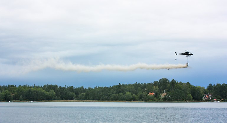 Fotografi på helikopter som sprider ut behandlad märgel i Kyrkviken. Ser ut som ett moln bakom helikoptern. 