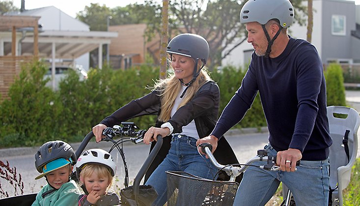 En kvinna och en man sittandes på varsin cykel. Kvinnan sitter på en lådcykel, och i lådan fram syns två glada barn. 