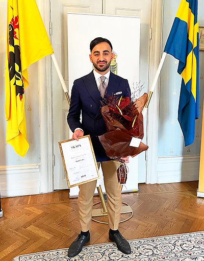 Muhib Lafta, vinnare av SKAPA:s länspris 2022, står i en salong, han håller i ett inglasat diplom och en stor bukett med blommor. Bredvid syns länsstyrelsens fana och Svenska flaggan. 