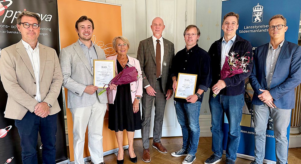 Sju människor står bredvid varandra i en sal. Diplom och blombuketter visas upp.