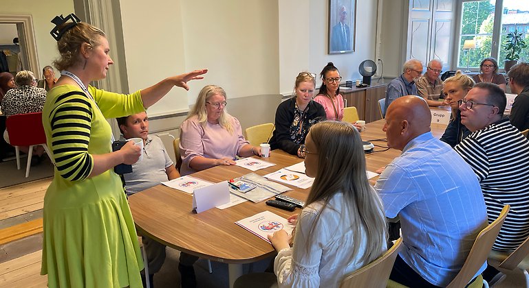 Ne kvinna står framför en samling personer. Deltagarna sitter runt ett bord och lyssnar.