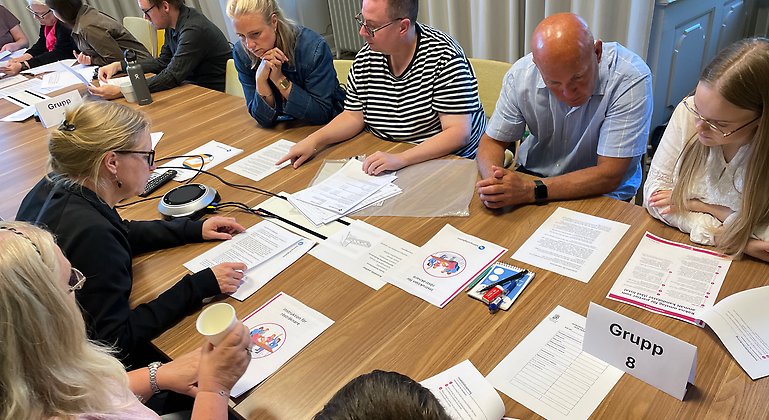 Ett större antal personer sitter runt ett bord. De tittar på instruktioner som ligger på bordet.