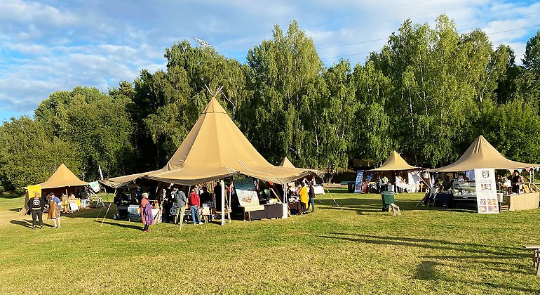 Matfestival med bruna tält, människor, gräsmatta, skog och en blå himmel. 