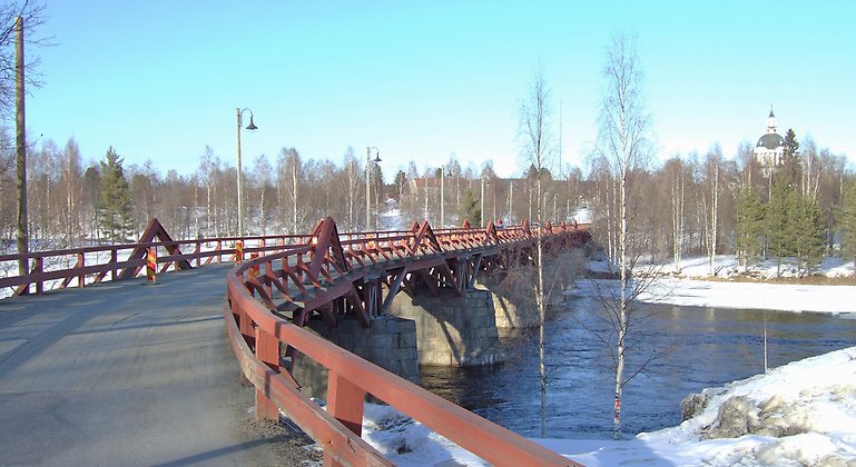 Röd träbro med pelare av stenmurverk som leder över älv. 