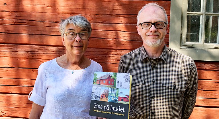 Anita Löfgren Ek och Patrick Björklund står mot en röd husvägg och håller upp ett exemplar av den nya skriften Hus på landet.